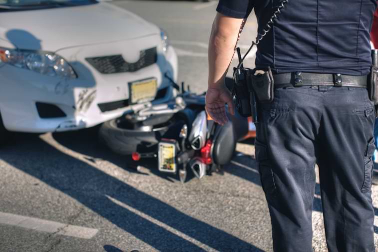 A police officer stands in front of a motorcycle underneath car after car and motorcycle accident 